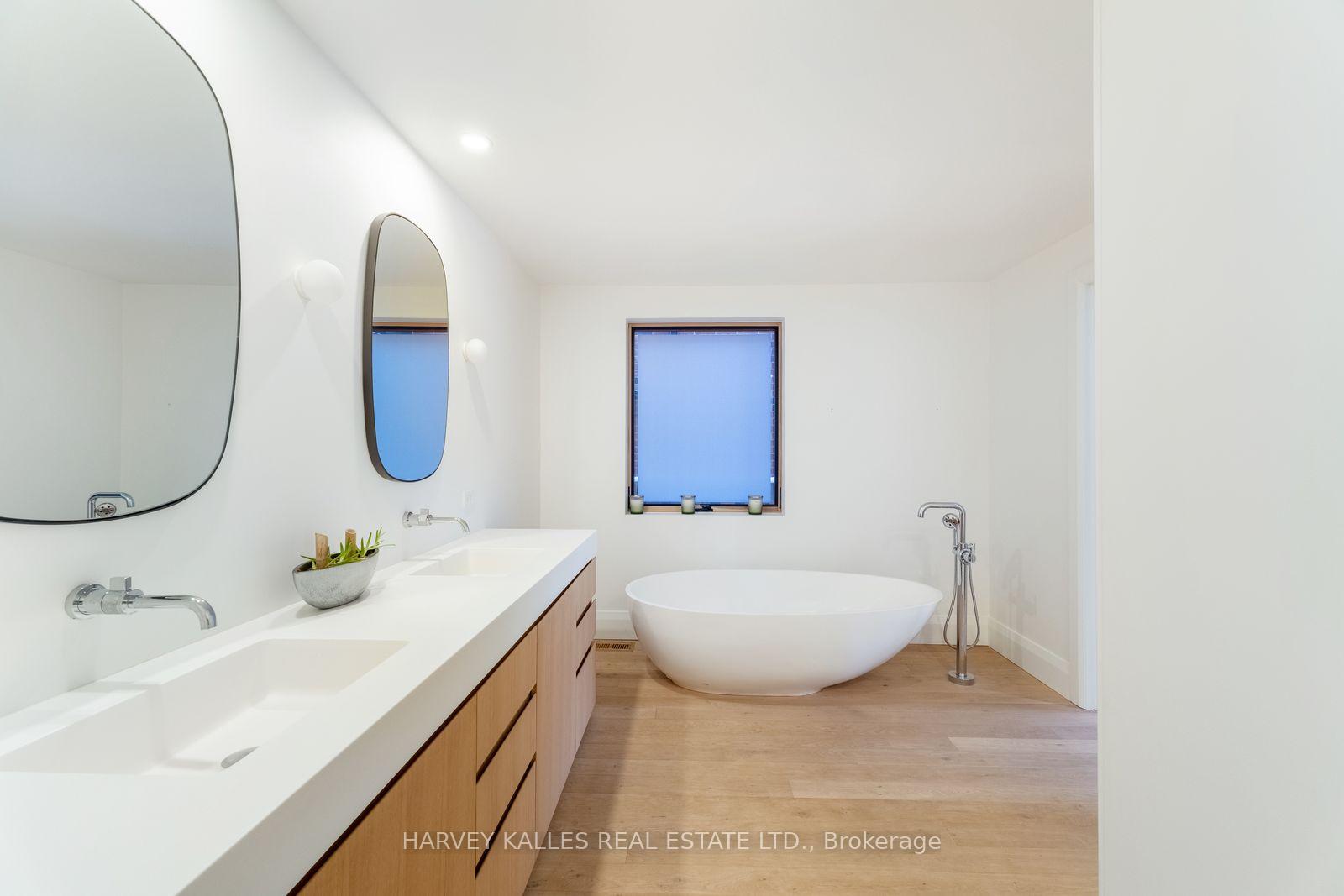 Bright bathroom with a double vanity, arched mirrors, and a freestanding tub beneath a window.