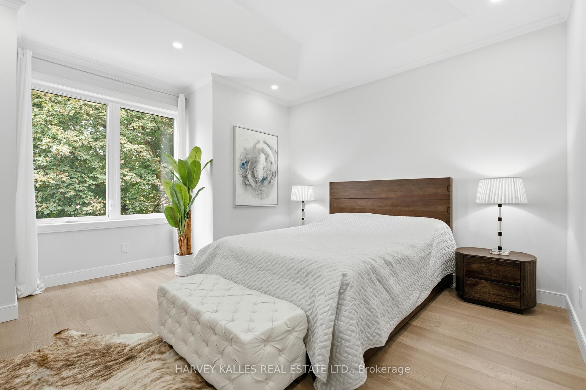 Bright bedroom with a wood-headboard bed, tufted bench, and a wide window overlooking leafy trees.