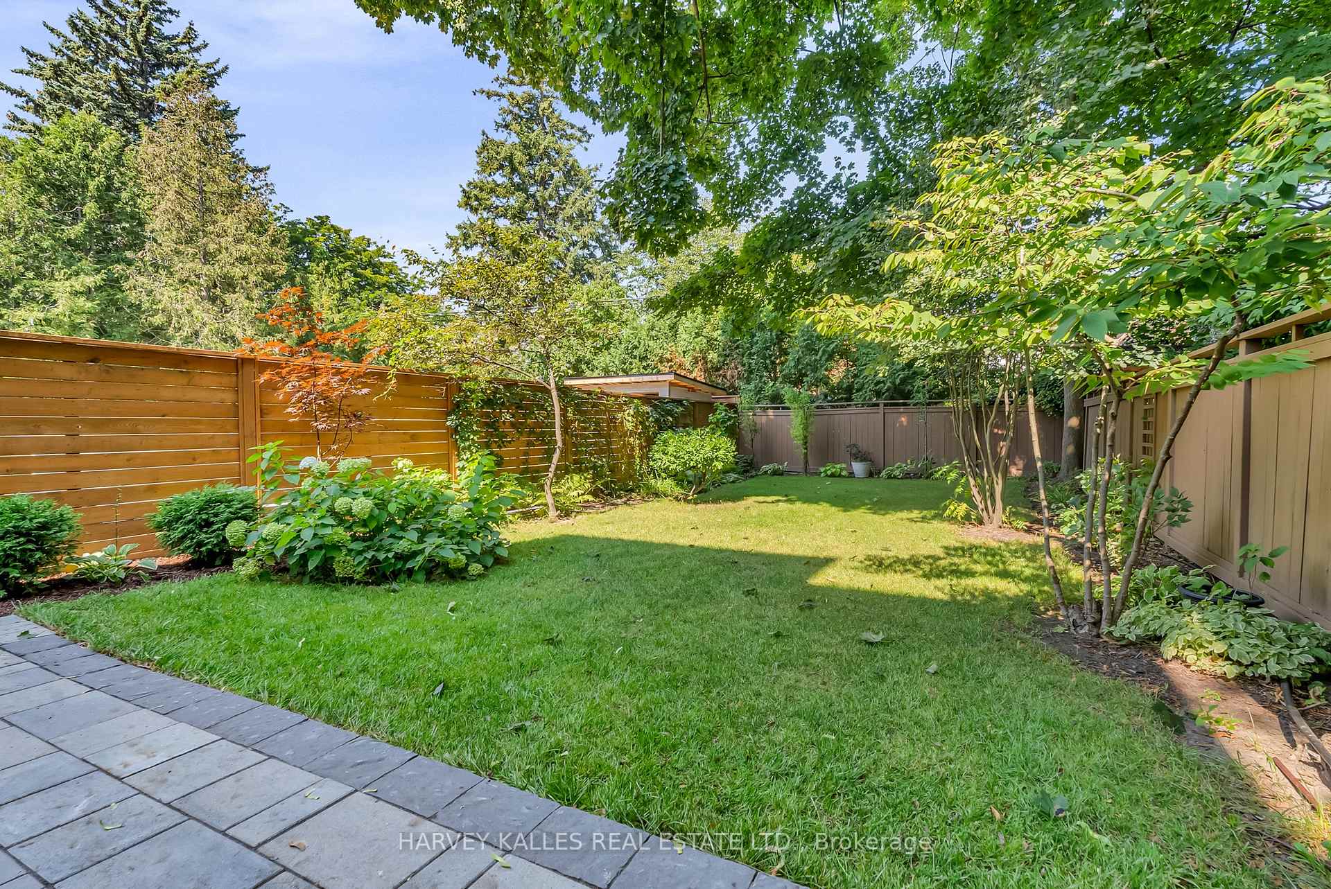 Landscaped backyard with a broad lawn, stone patio, wooden privacy fences, garden beds, and mature trees.