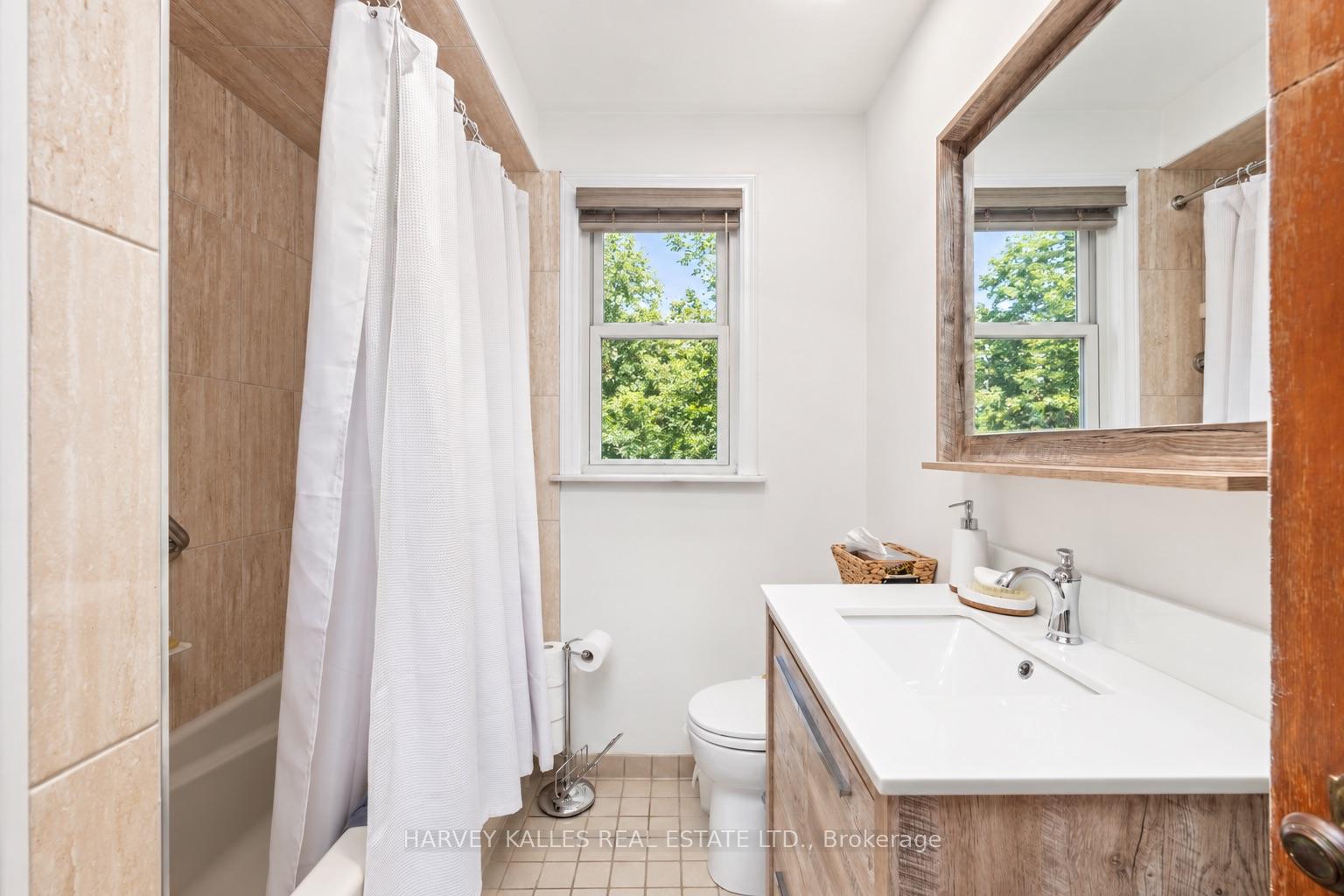 Bathroom with a wood-finish vanity, beige-tiled tub and shower, and a window overlooking leafy trees.
