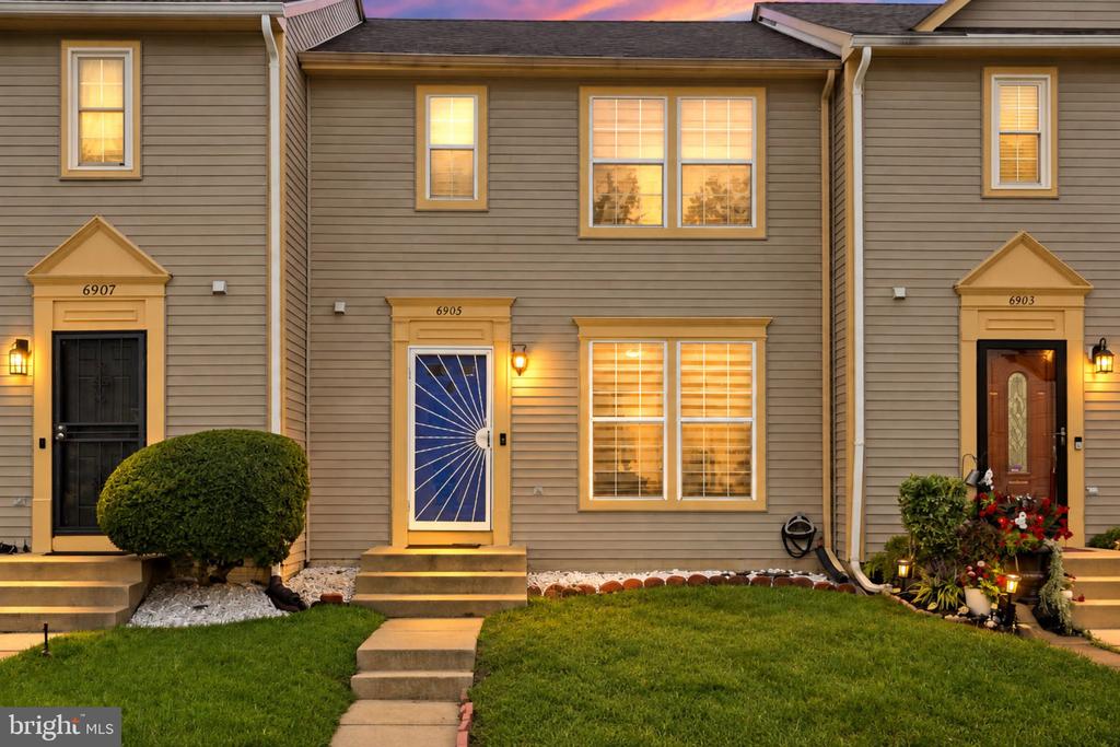 Townhouse exterior at dusk with a blue front door, lit windows, and a walkway across the lawn.