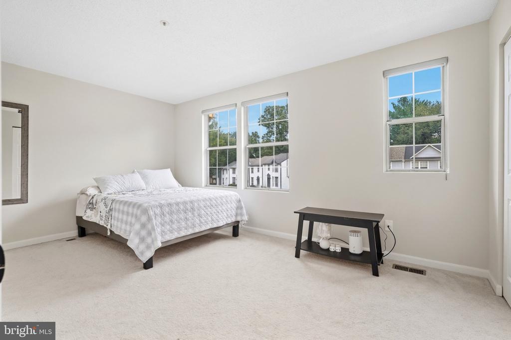 Bright bedroom with a white bed, light carpeting, and three windows overlooking neighboring homes.