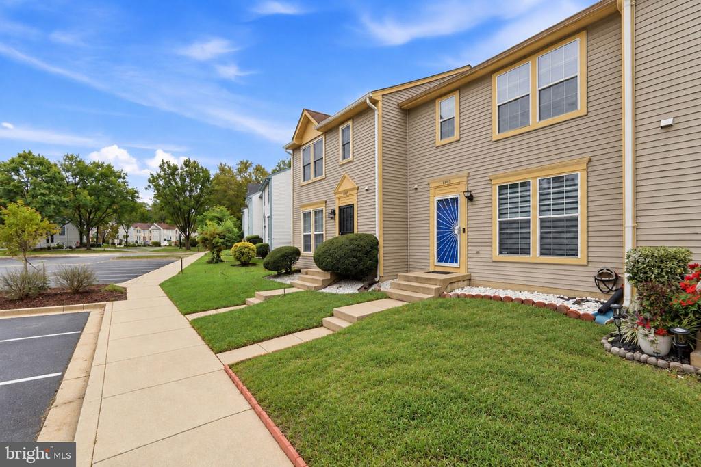 Attached tan townhomes with yellow trim, a blue front door, and landscaped lawns beside a sidewalk.