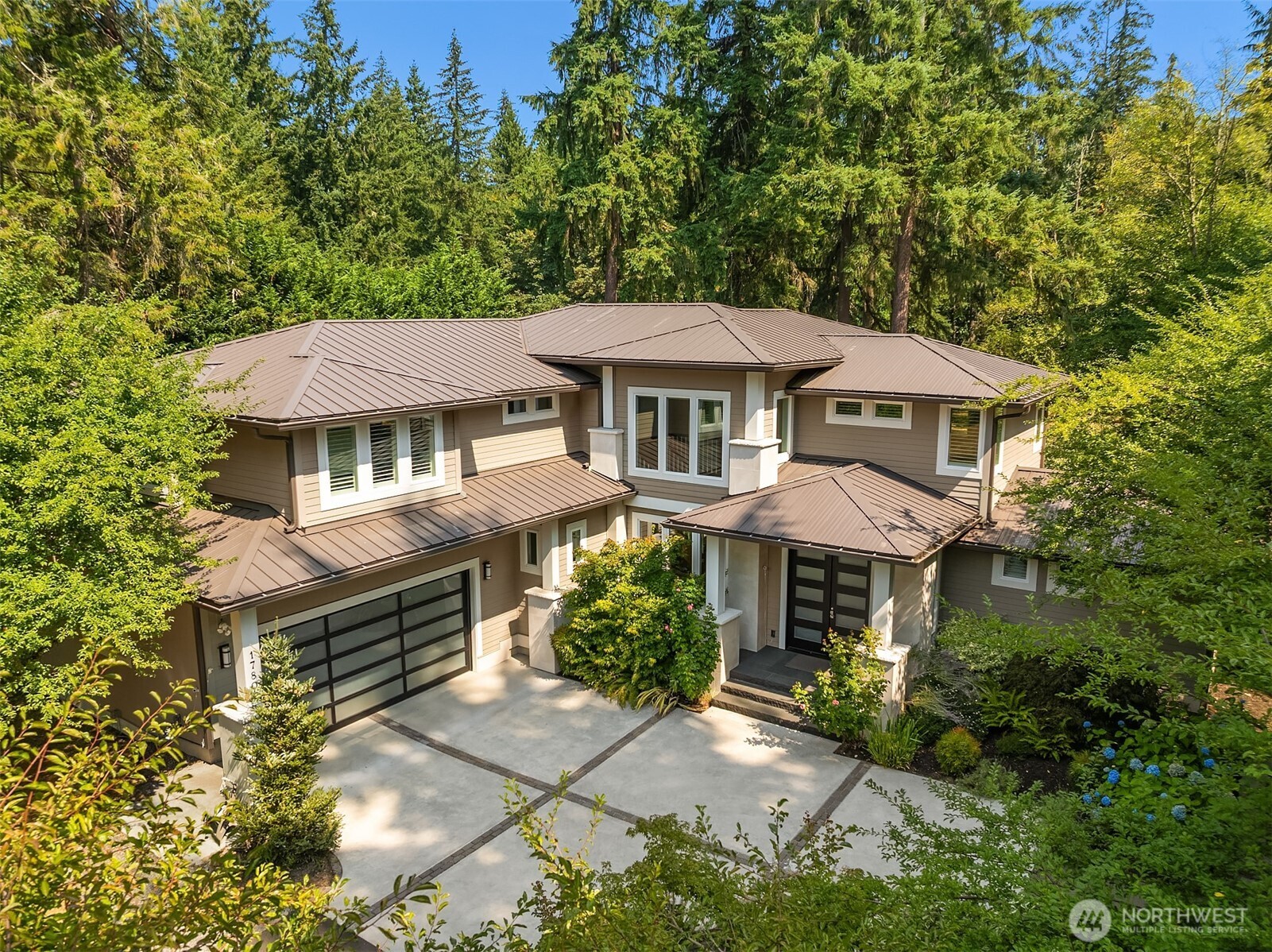 Contemporary two-story home with a broad metal roof, landscaped driveway, and dense evergreen surroundings.