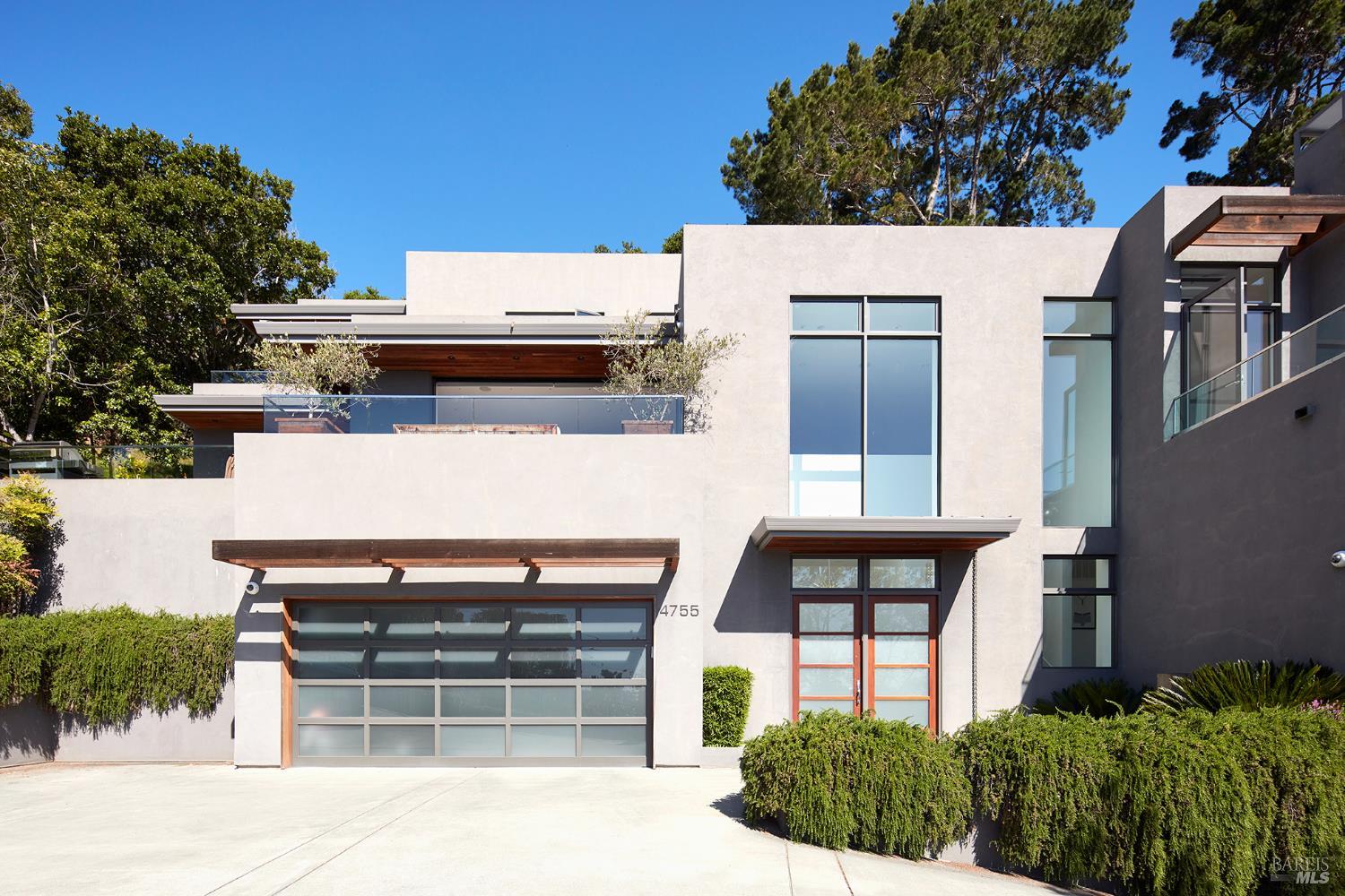 Modern multi-level home with a glass garage door, tall windows, and landscaped hedges.