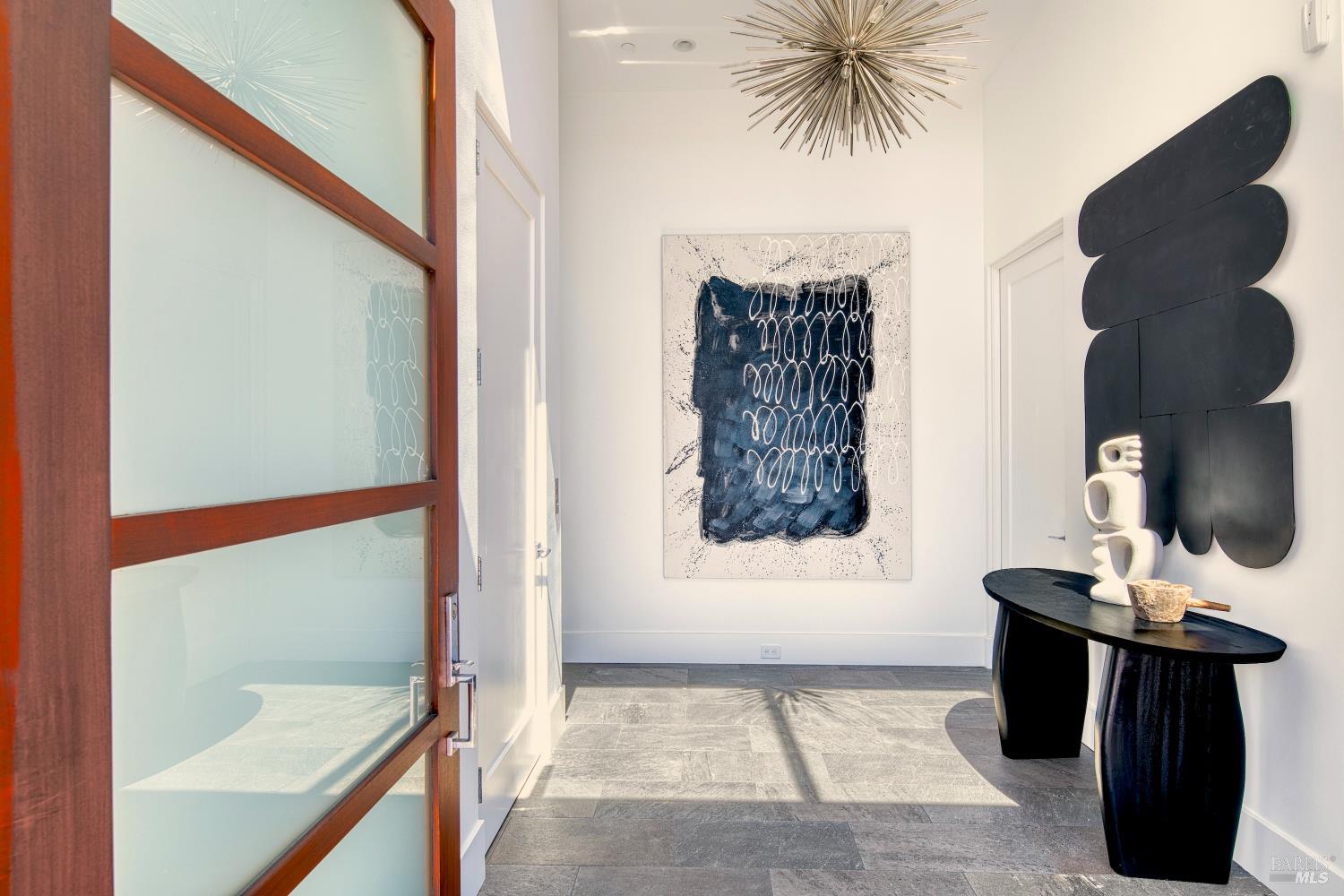 Furnished entry foyer with a wood-framed glass door, abstract artwork, sculptural console, and starburst chandelier.