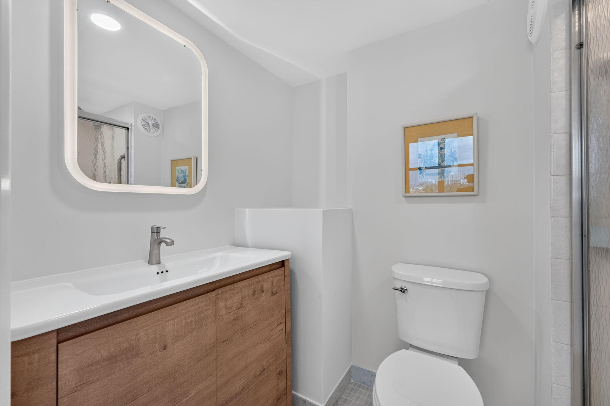 Modern bathroom with a wood-front vanity, illuminated mirror, toilet, and glass-enclosed shower.