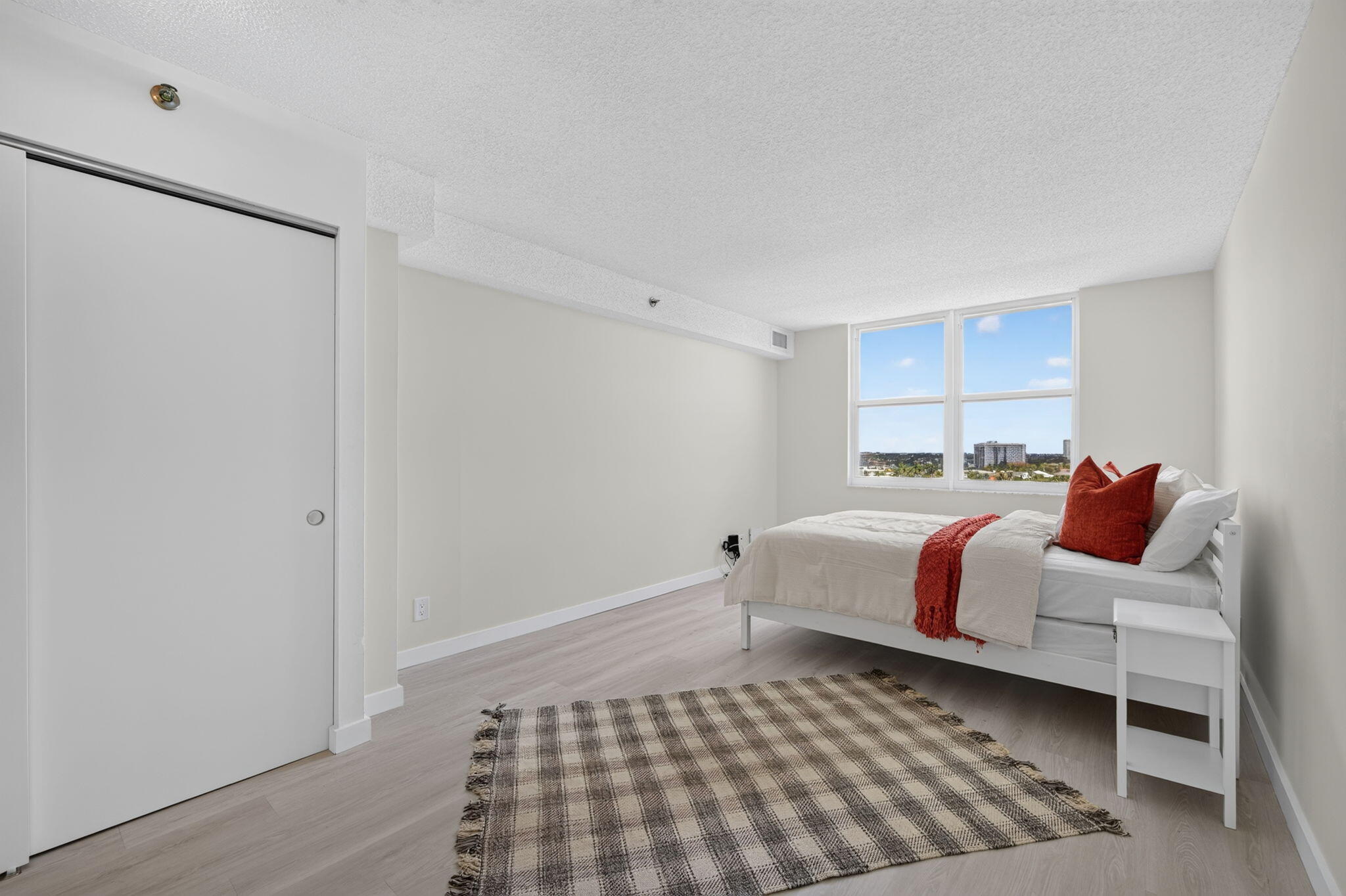 Staged bedroom with a white bed, rust-red accents, plaid rug, and windows overlooking nearby buildings.