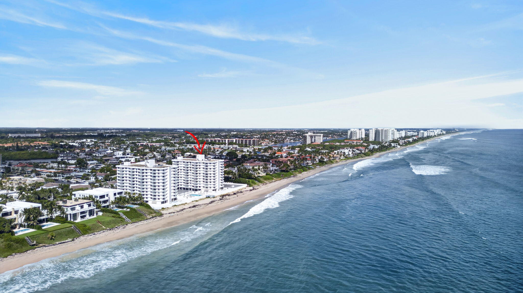 Aerial view of an oceanfront condominium marked by a red arrow, with the beach and coastline stretching into the distance.