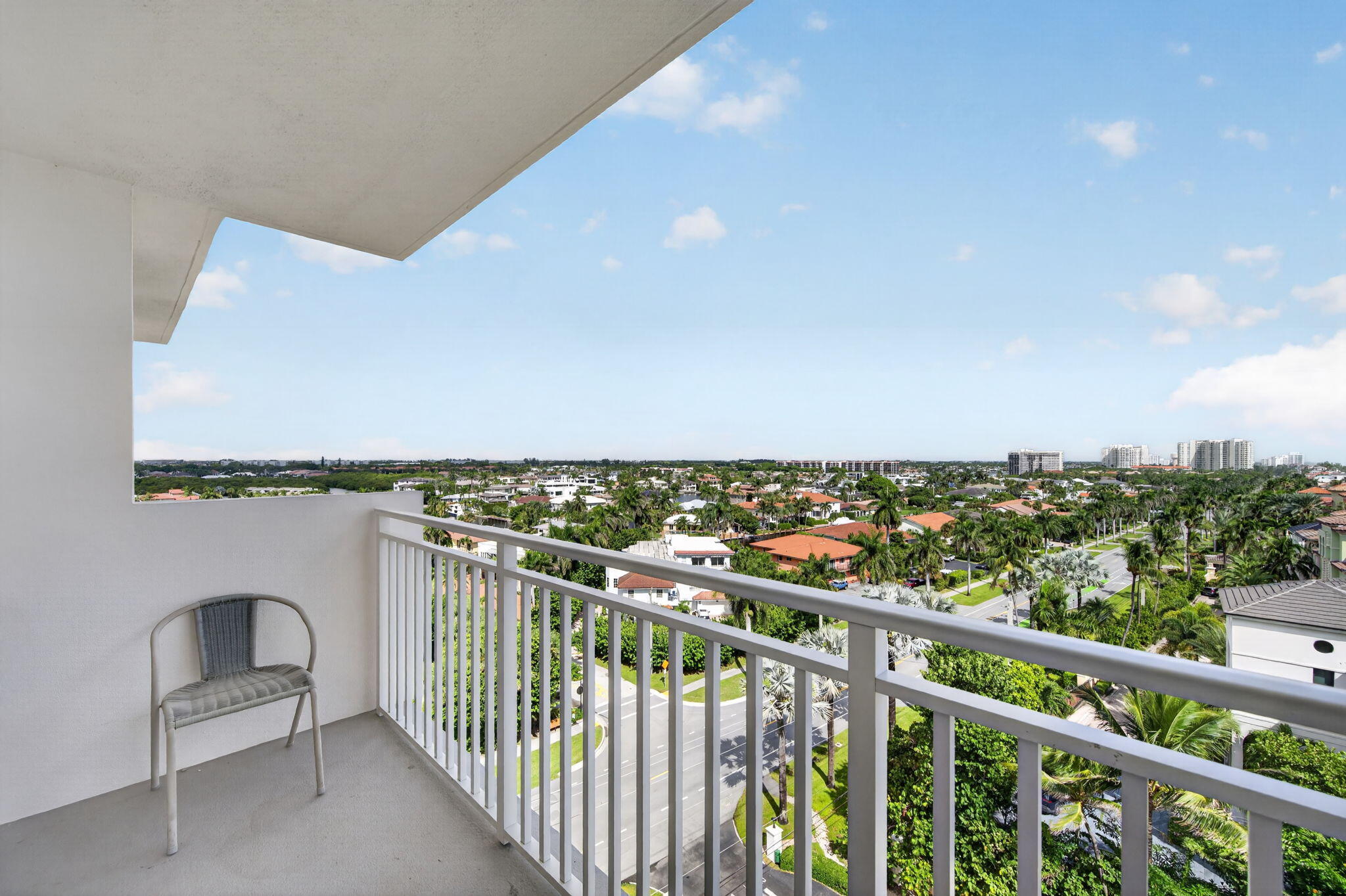 High-rise balcony overlooking a palm-lined neighborhood, with a white railing and distant condominium buildings.