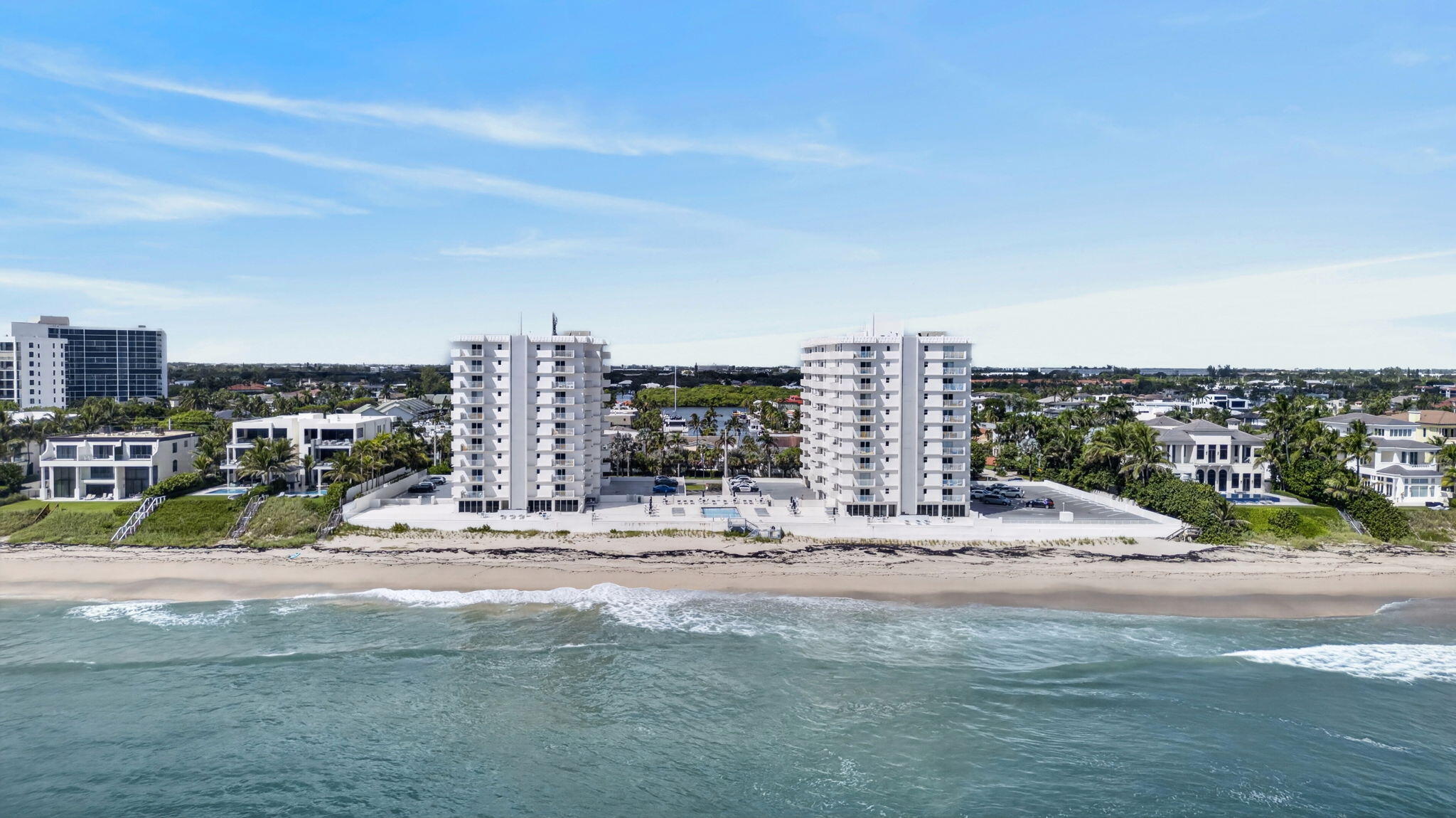 Two white condominium towers stand beside a sandy beach, viewed from over the ocean.