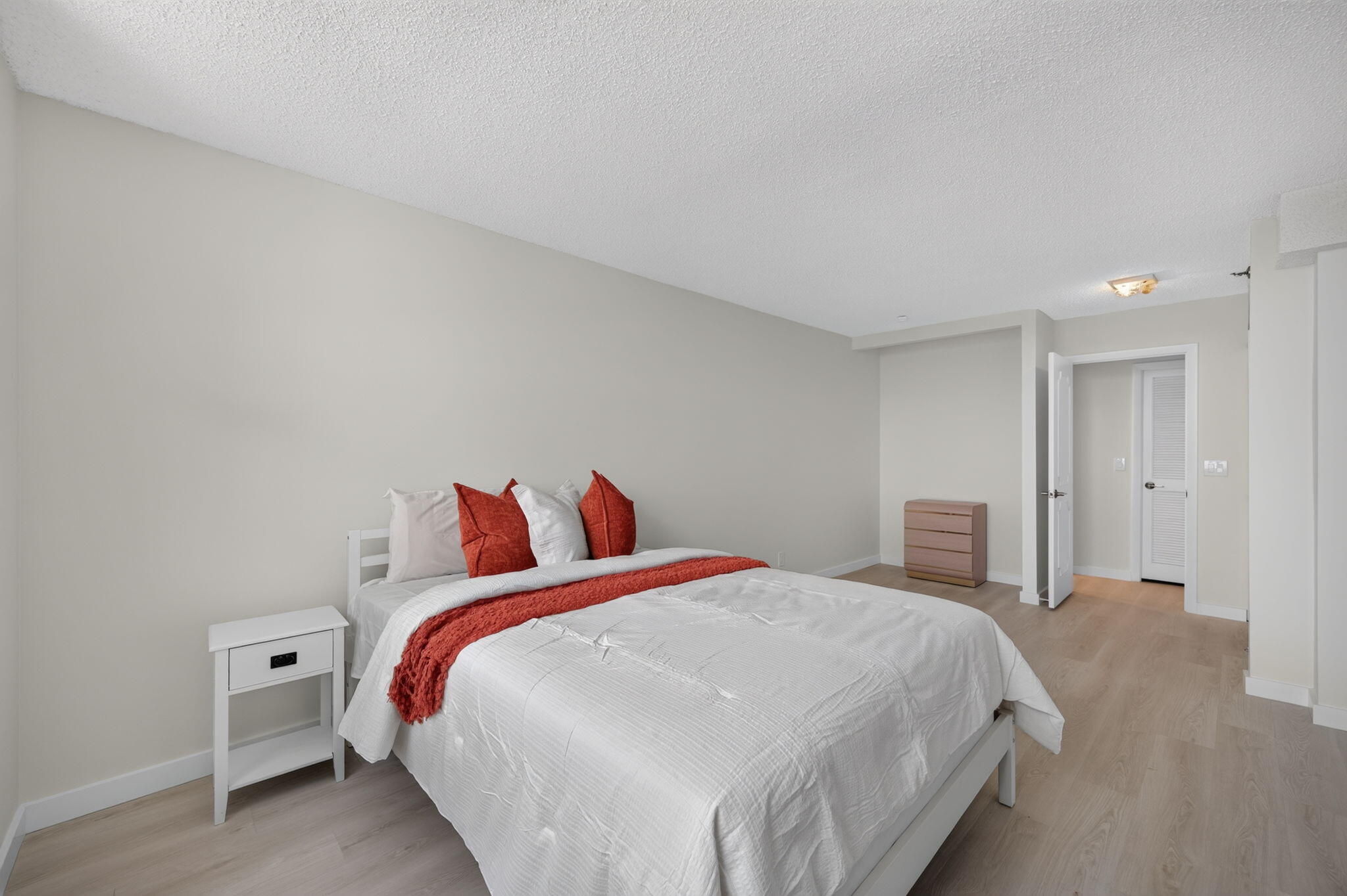 Bedroom with a white bed, rust-red accents, pale wood floors, and an open doorway.