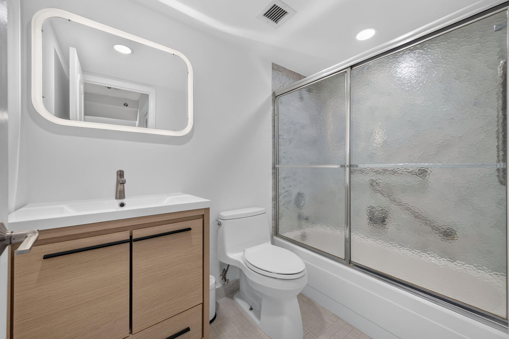 Bathroom with a light wood vanity, white sink, toilet, and textured glass shower doors.