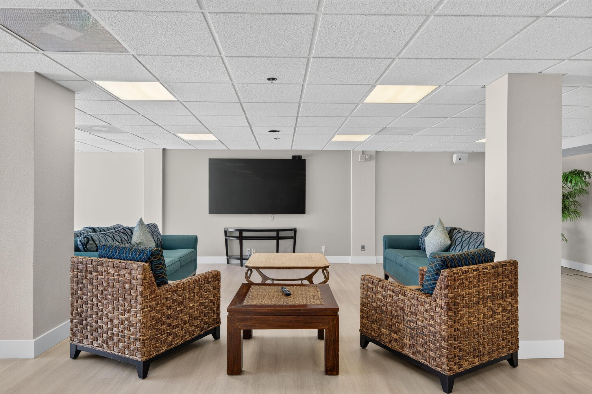 Furnished lounge with teal sofas, woven wicker chairs, and a wall-mounted television.