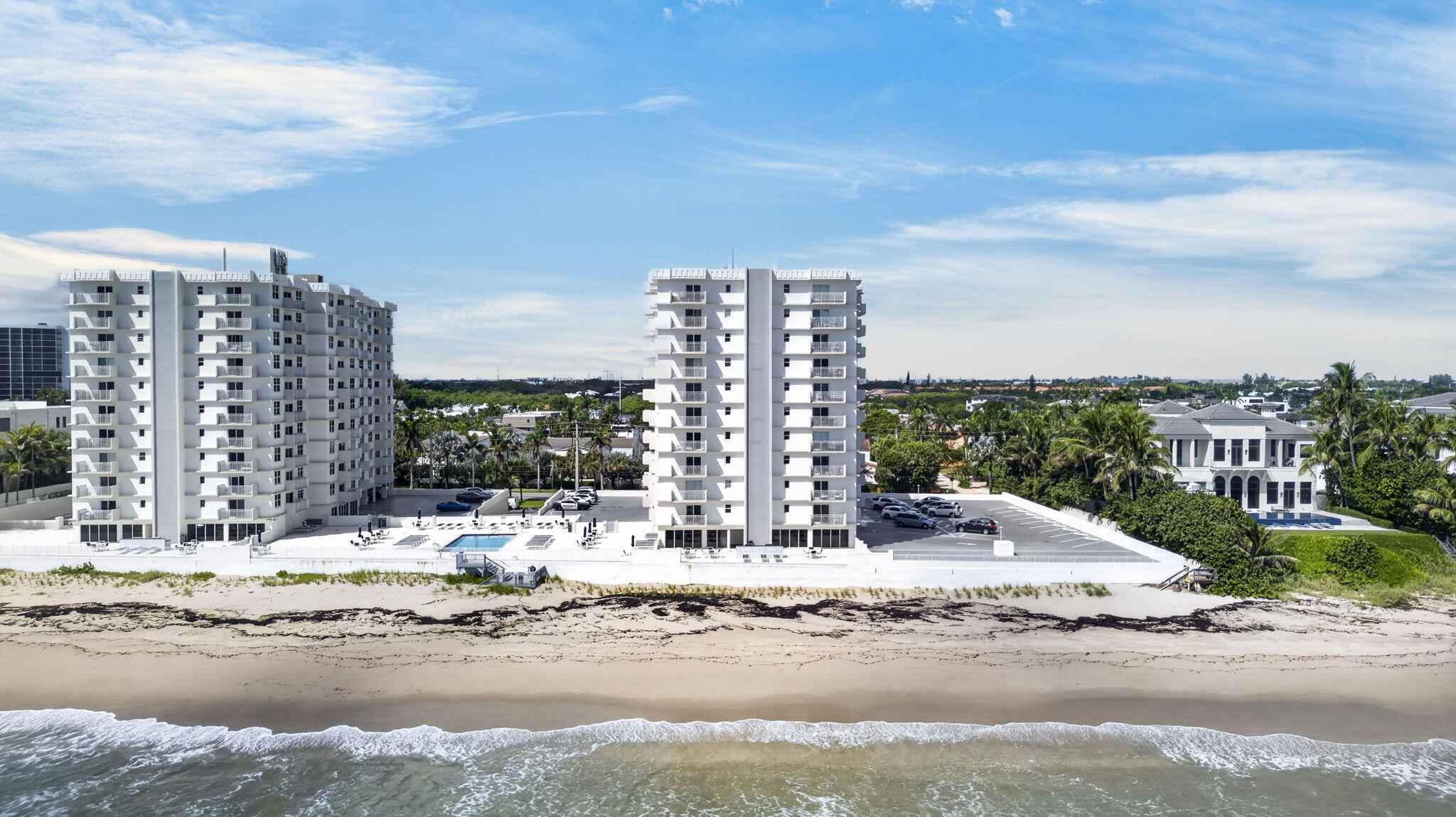 Two white beachfront condominium buildings beside a sandy beach, with a pool deck and oceanfront view.