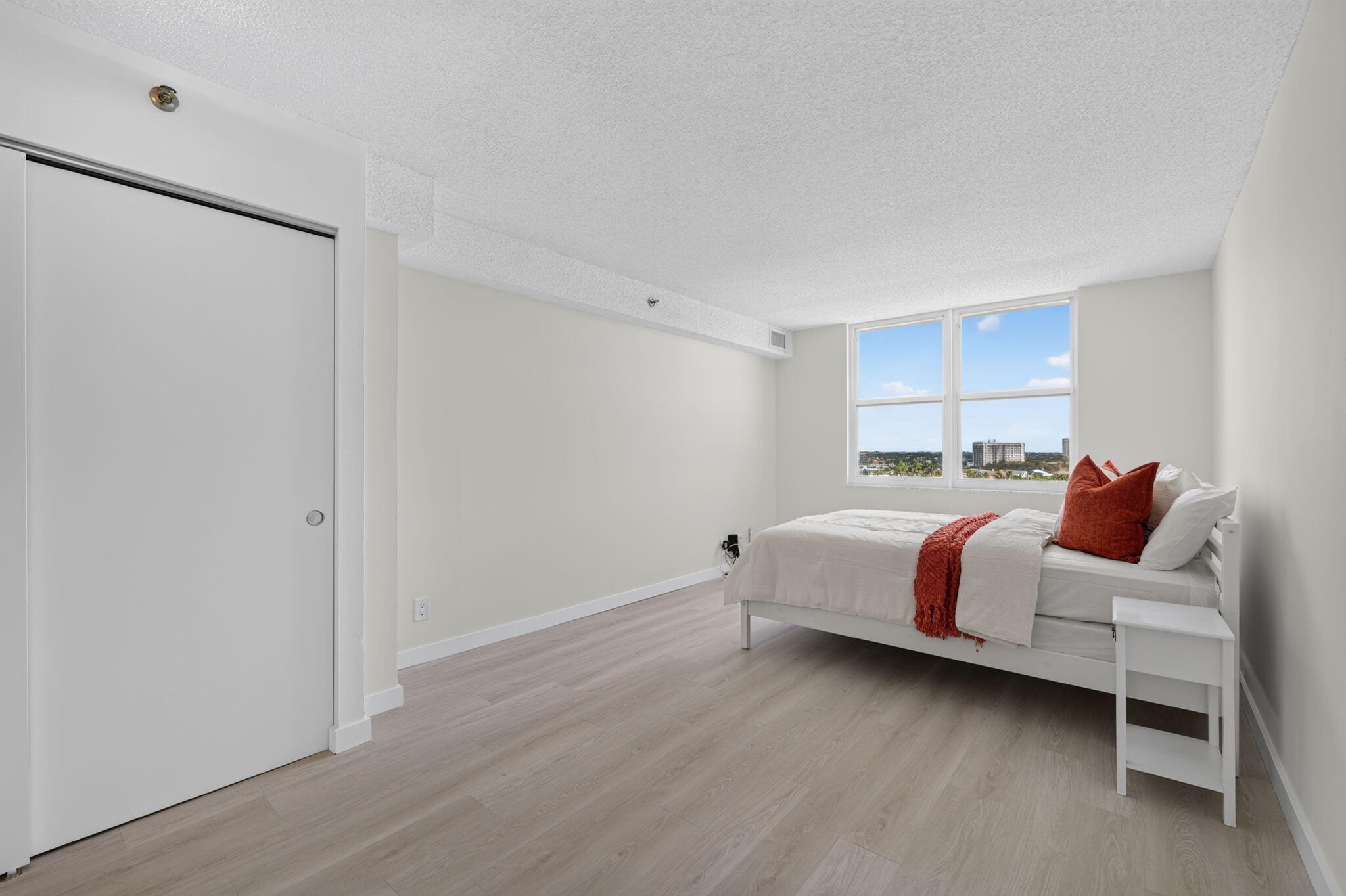 Bedroom with a white bed, rust-red accents, light wood flooring, and two large windows.