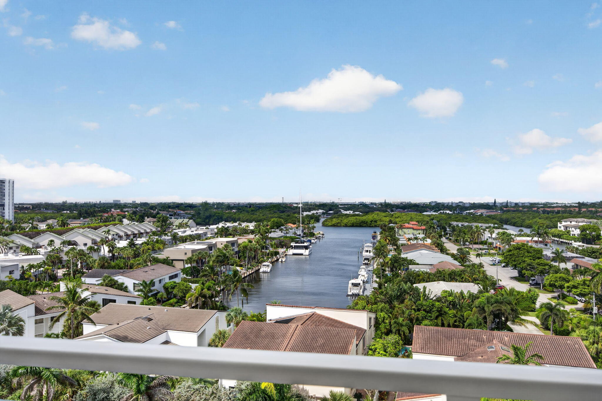 Residential canal with moored boats and palm-lined homes, viewed over a balcony railing.