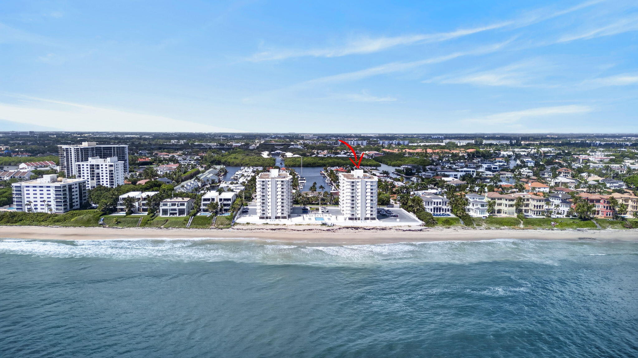 Aerial view of a beachfront condominium marked by a red arrow, with ocean and marina nearby.