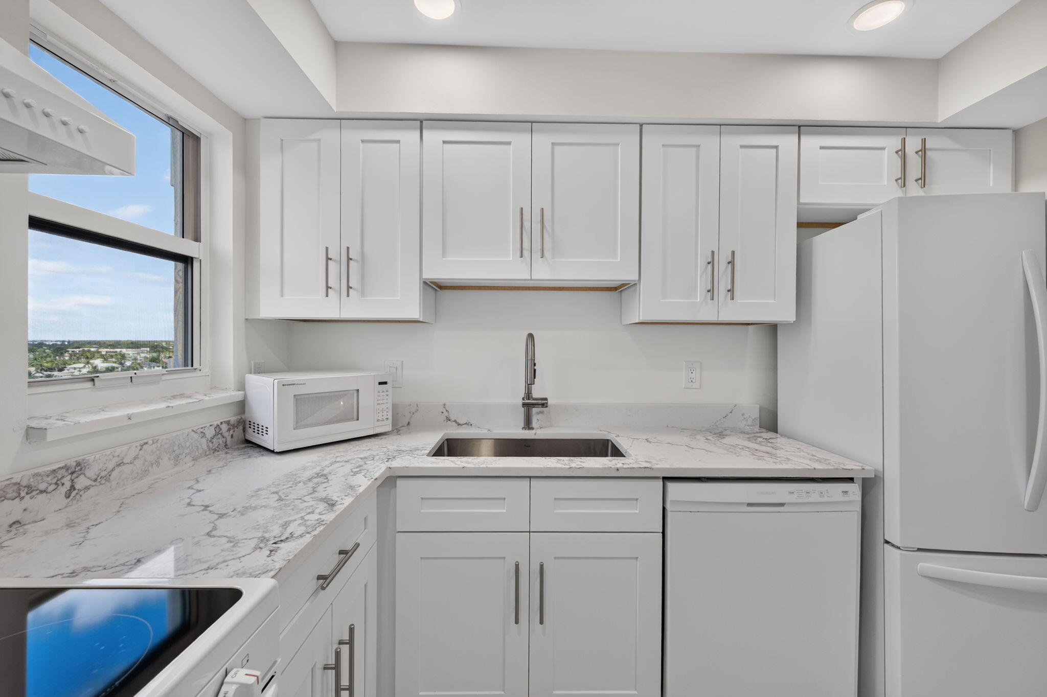 Bright white kitchen with marble-look counters, a large window, and modern appliances.