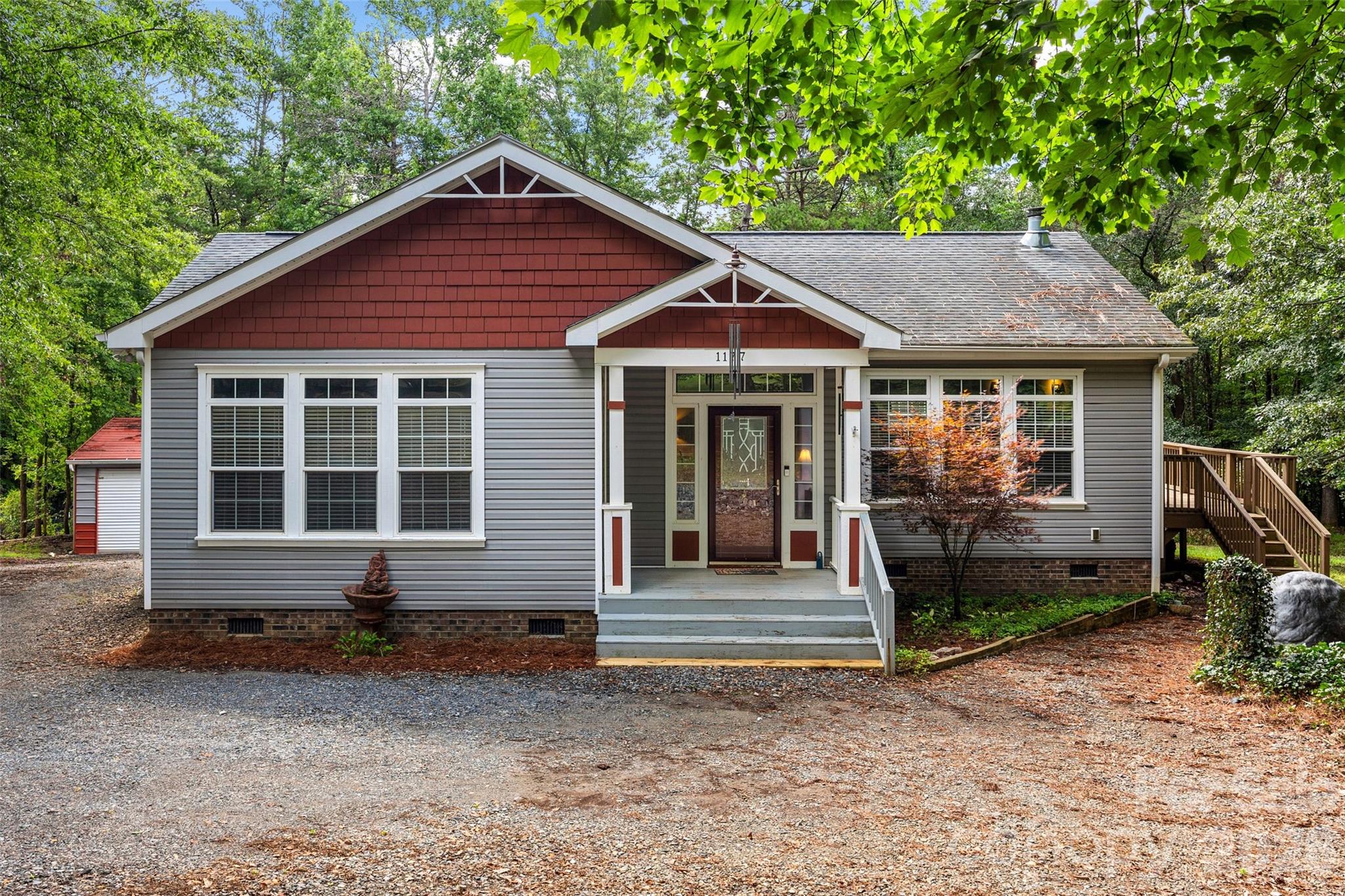 Gray-sided single-story house with red gables, a covered porch, and gravel driveway beneath leafy trees.