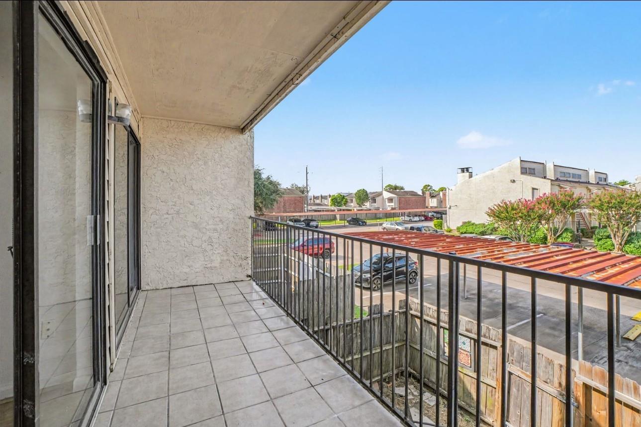 Covered tiled balcony overlooking a parking lot, with sliding glass doors and a black metal railing.