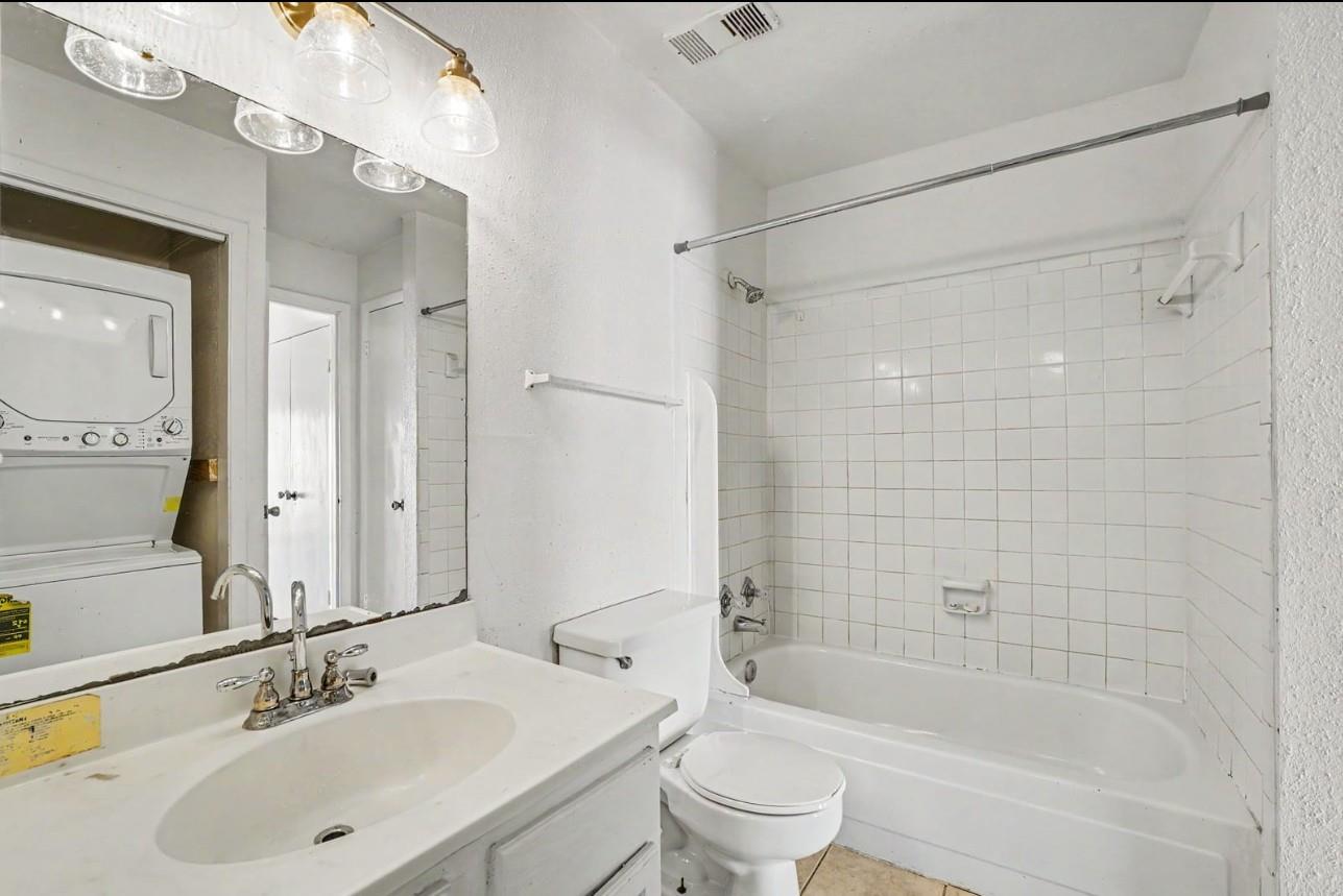 Bathroom with a white vanity, tiled tub-shower, toilet, and stacked washer and dryer reflected in the mirror.