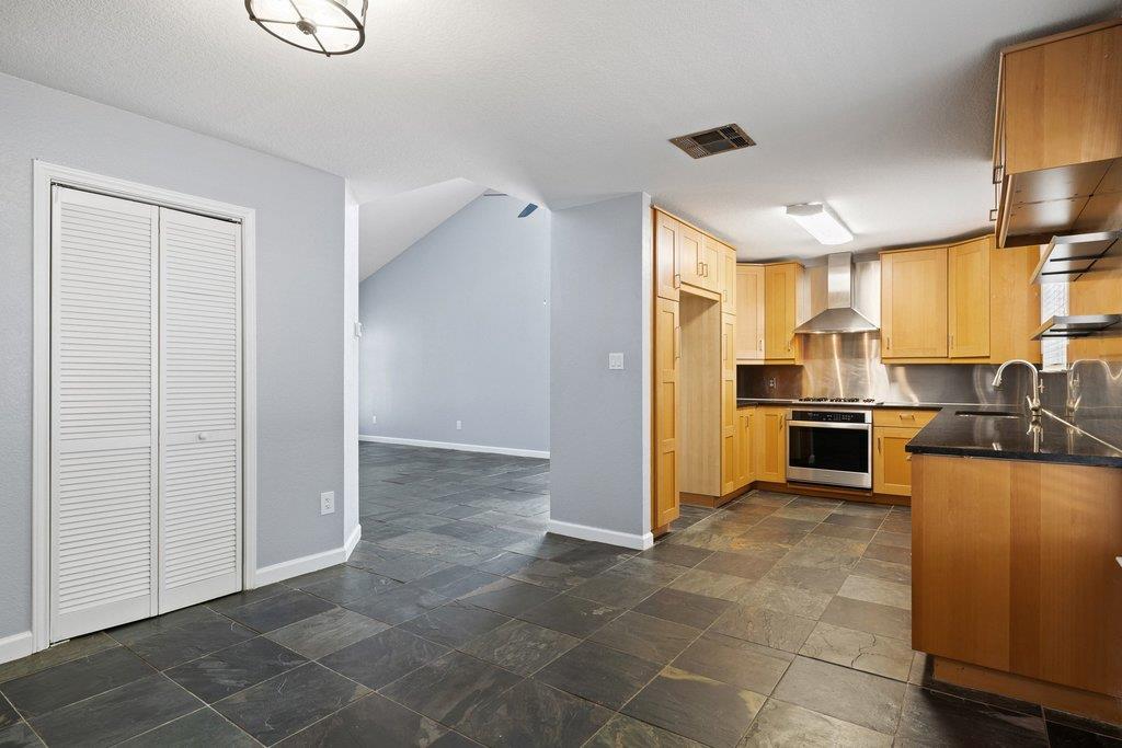 Vacant kitchen with wood cabinets, stainless steel appliances, and dark tile flooring opening to an adjoining room.