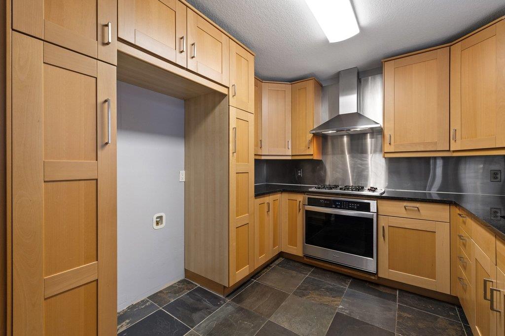 Kitchen with light wood cabinetry, stainless steel range hood, and dark tile flooring.
