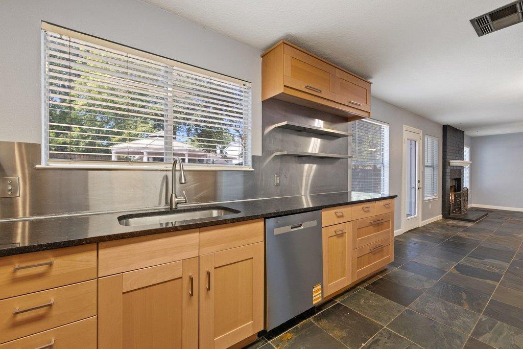 Kitchen with light wood cabinetry, dark countertops, stainless-steel backsplash, and large windows with blinds.