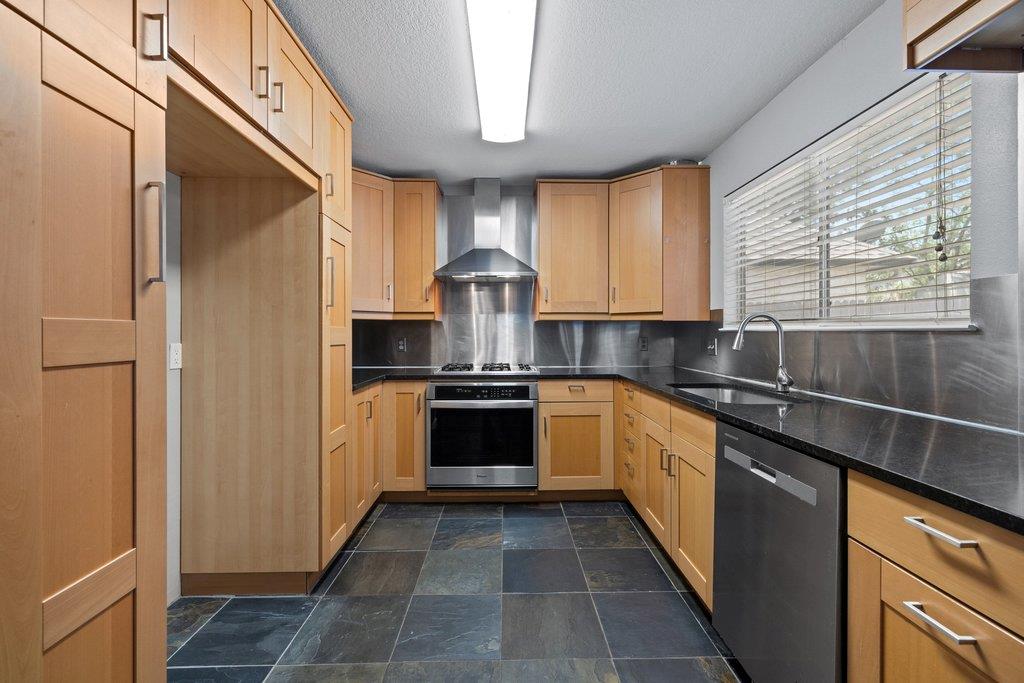 Kitchen with light wood cabinets, dark countertops, and a stainless steel range beneath a metal hood.