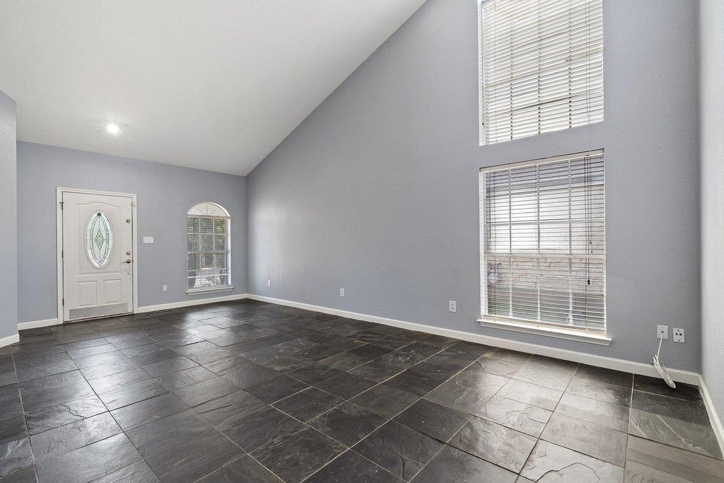 Vacant living room with dark tile floors, a vaulted ceiling, and tall windows with blinds.