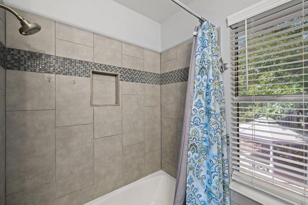 Bathroom tub and tiled shower with a blue patterned curtain beside a window with blinds.