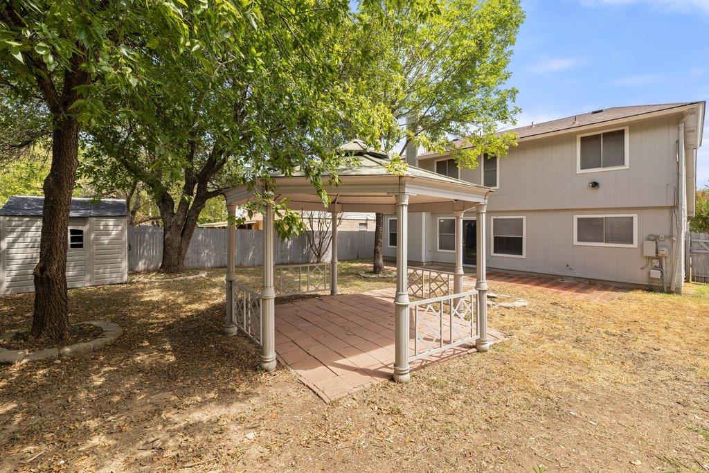 Two-story home behind a covered gazebo and paved patio, shaded by mature trees.