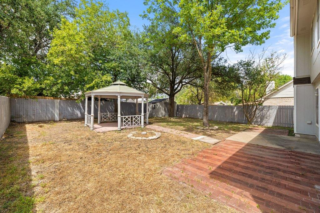 Fenced backyard with a white gazebo, mature trees, dry lawn, and brick patio.