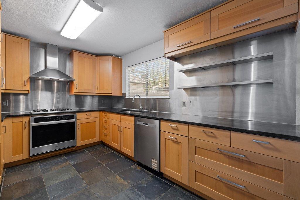 Kitchen with wood cabinetry, stainless steel appliances, black countertops, and dark tile flooring.