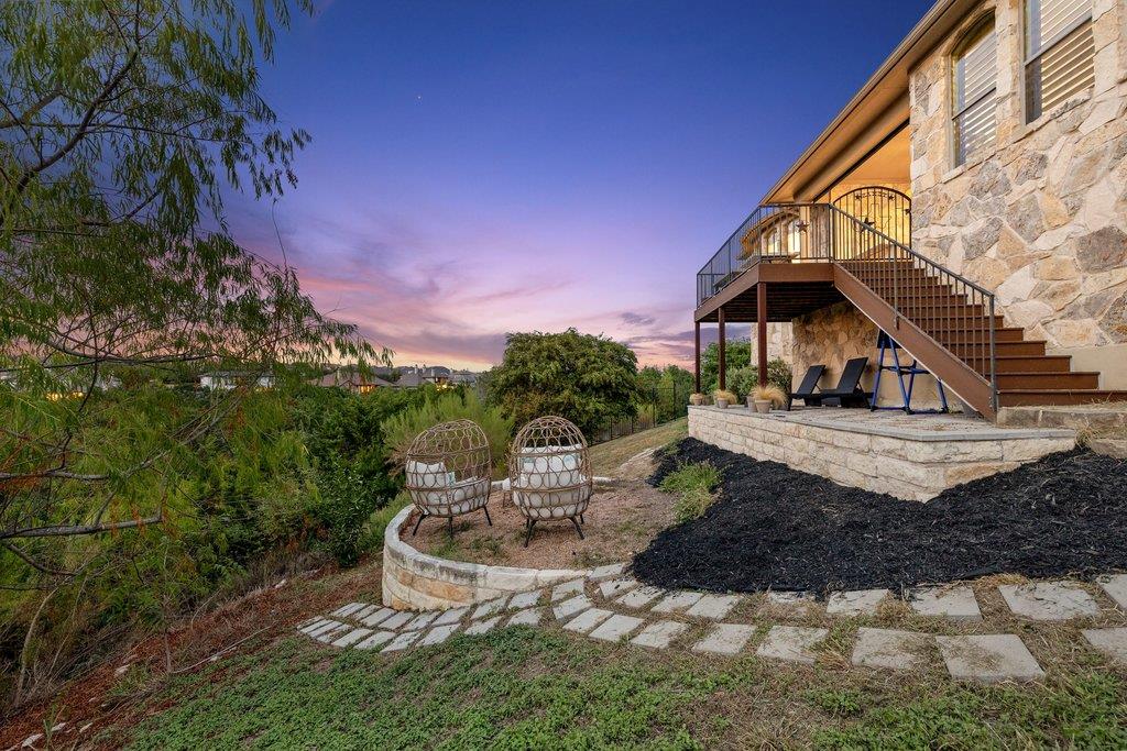 Stone home backyard with raised deck, outdoor chairs, curved planter, and paved path at dusk.