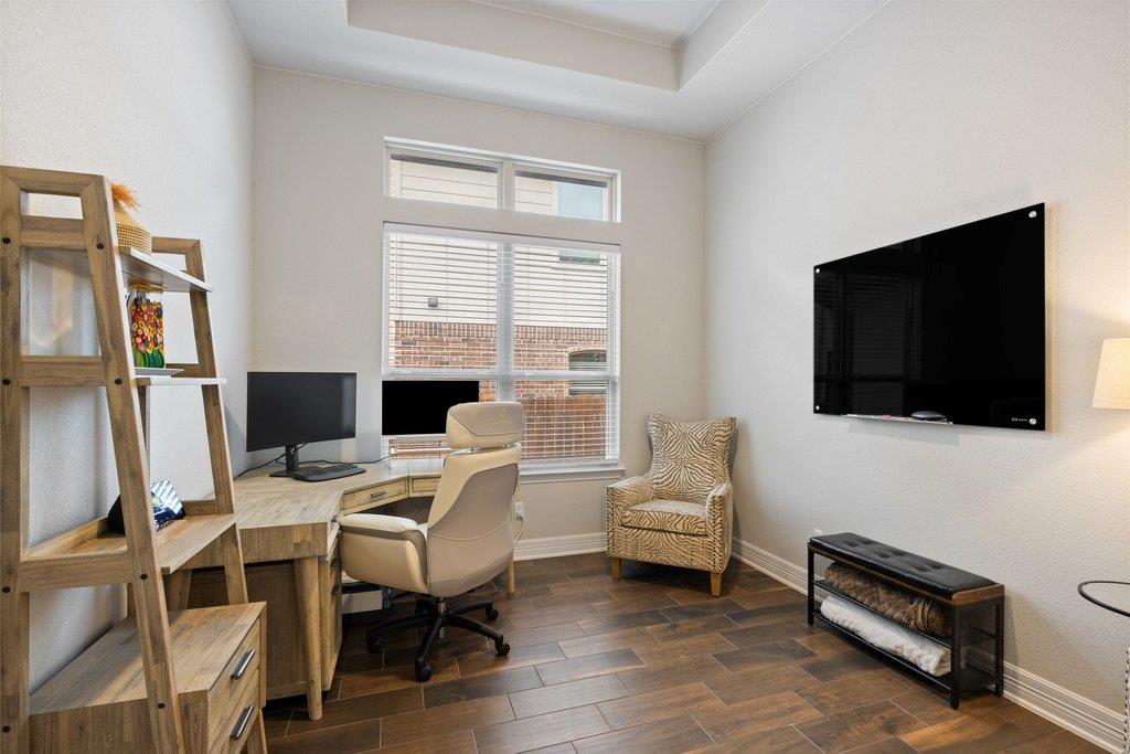 Home office with a wood desk beneath tall windows and a wall-mounted television.