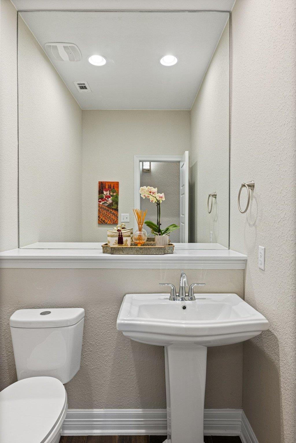 Bathroom with a white pedestal sink, toilet, large mirror, and orchid arrangement.