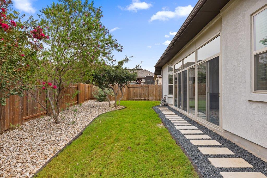 Fenced backyard with a green lawn, flowering trees, and stepping stones beside the house.