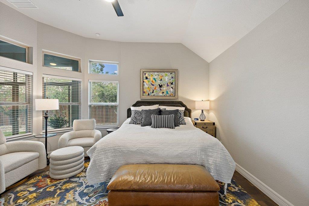 Furnished bedroom with a white-quilted bed, tall windows, cream armchairs, and a colorful patterned rug.