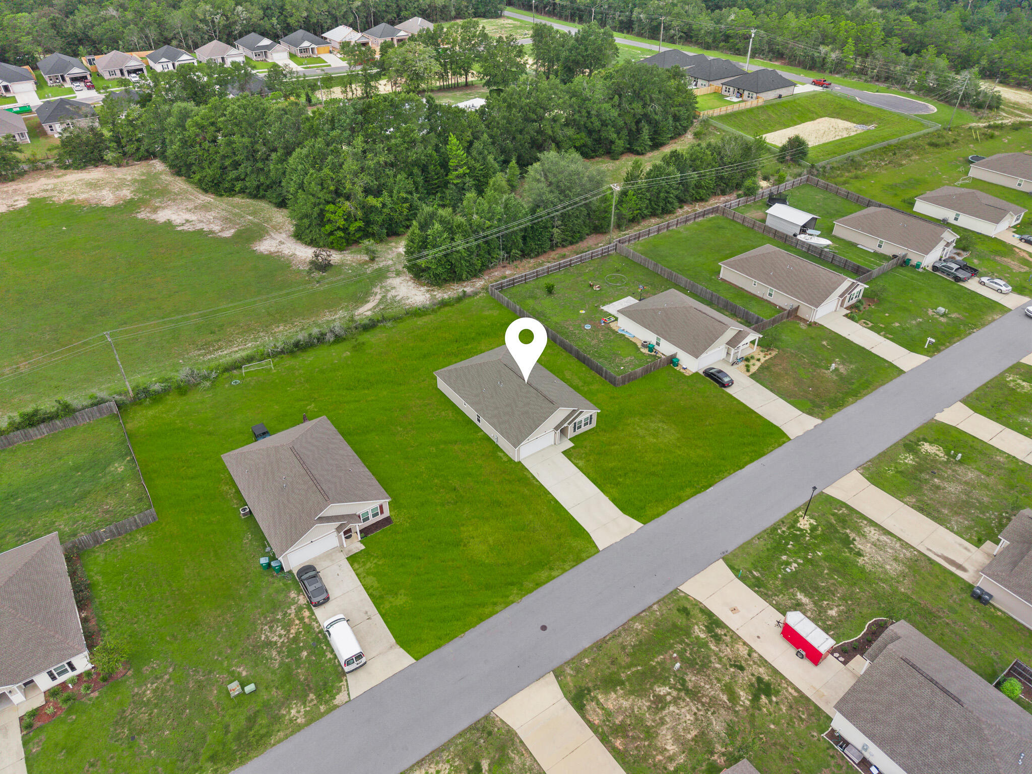 Aerial view of a single-story home on a broad lawn, marked by a white location pin beside the road.