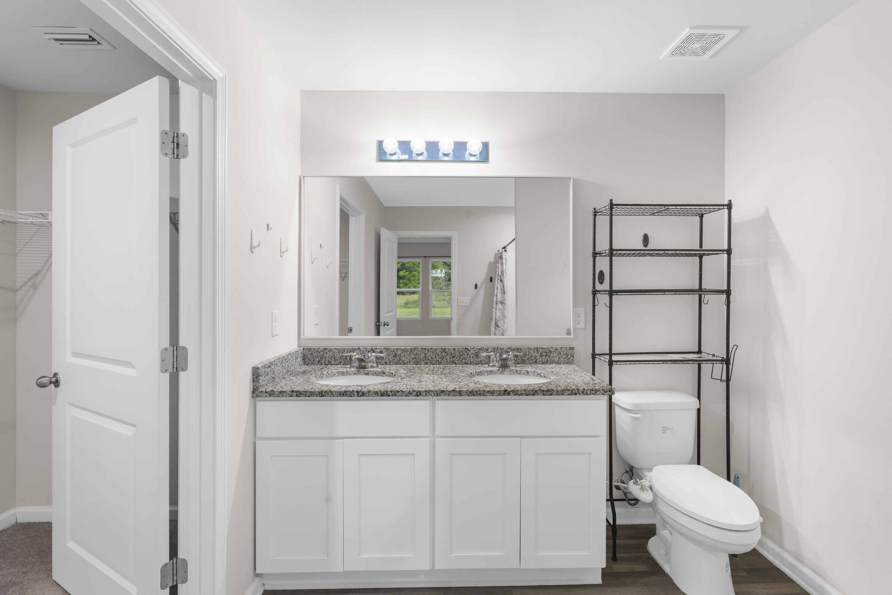 Bathroom with a double-sink granite vanity and toilet beside black metal shelving.