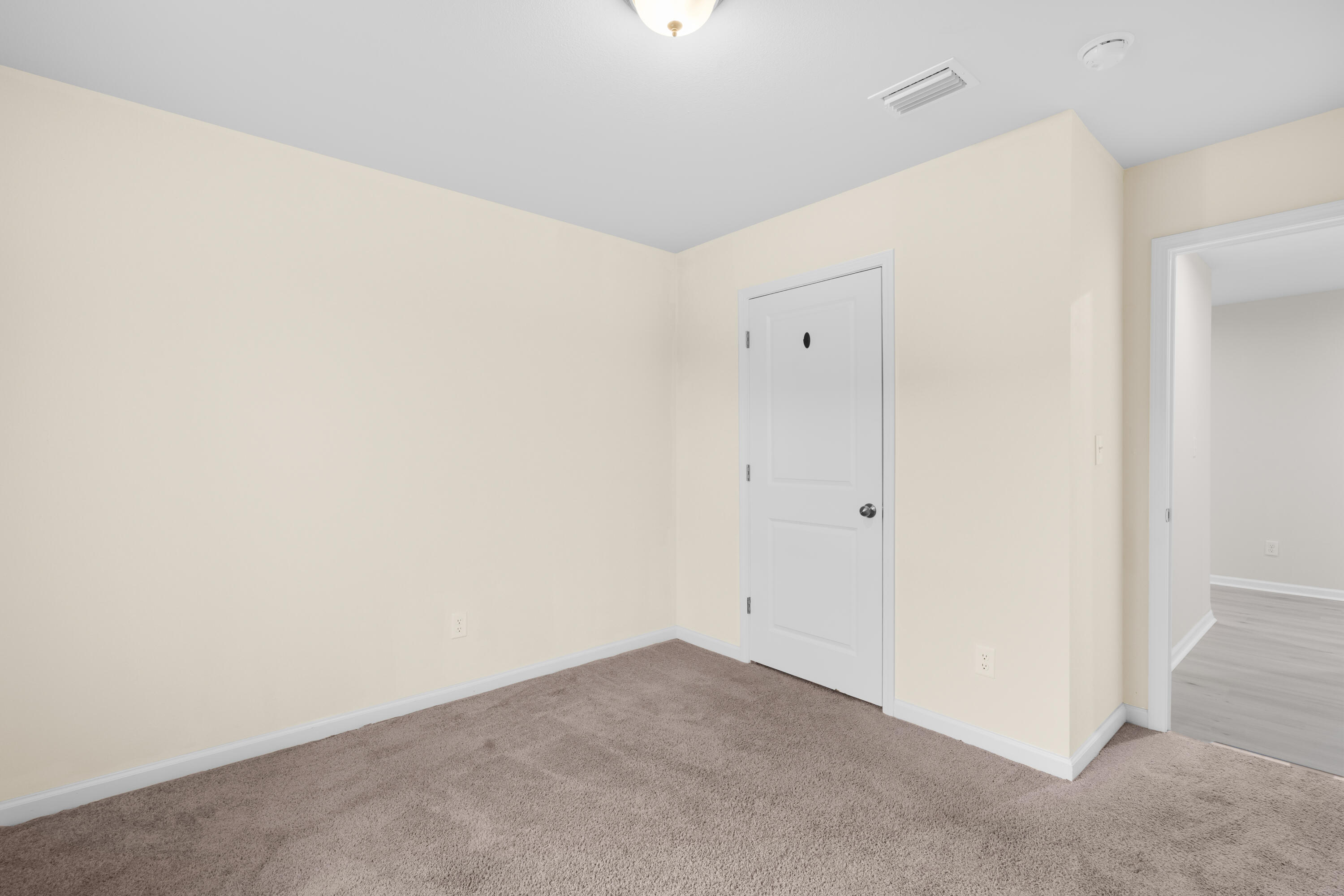 Empty carpeted room with pale walls, a closed door, and an open doorway to a wood-look floor.