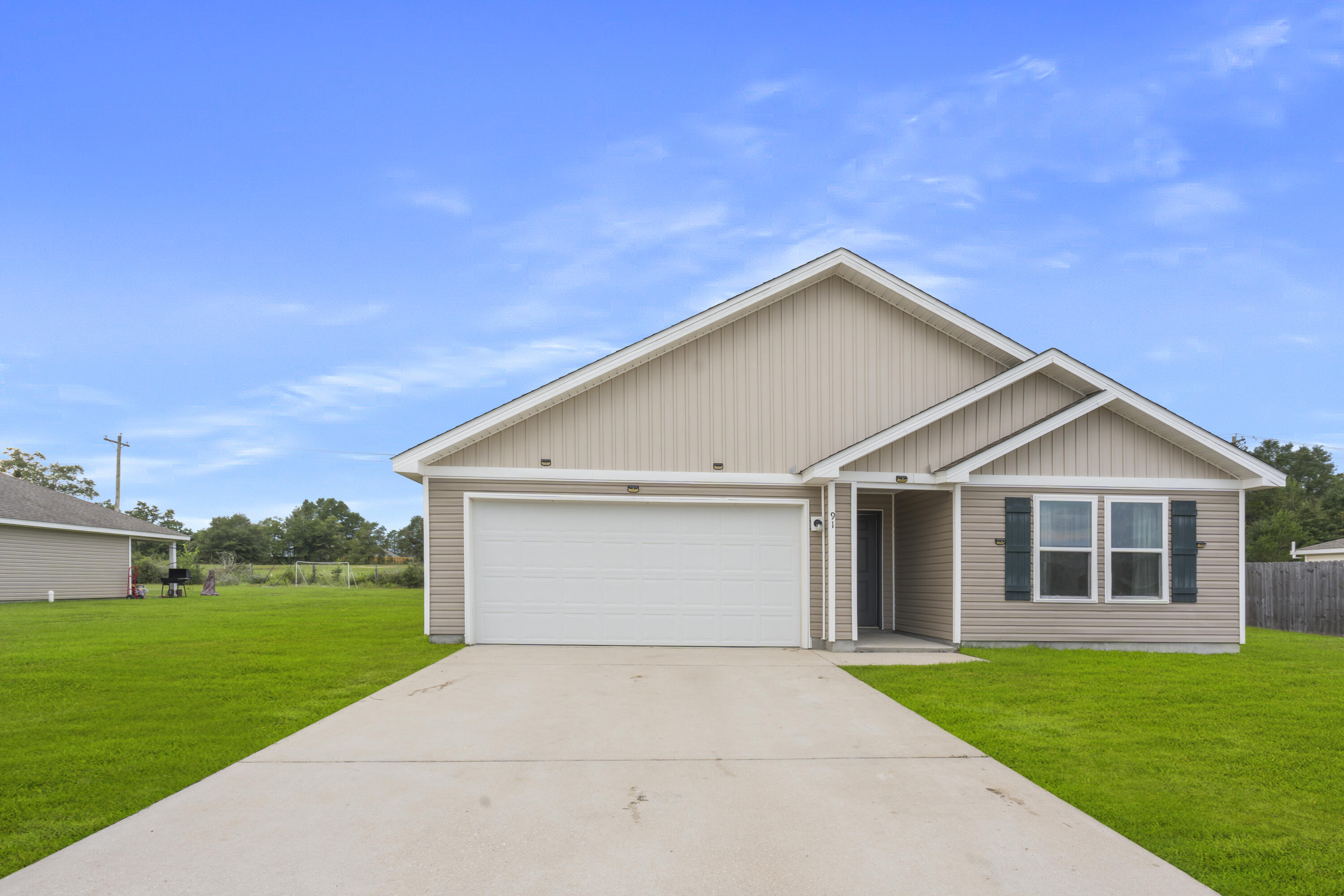 Beige single-story home with a two-car garage, dark shutters, and a wide concrete driveway.