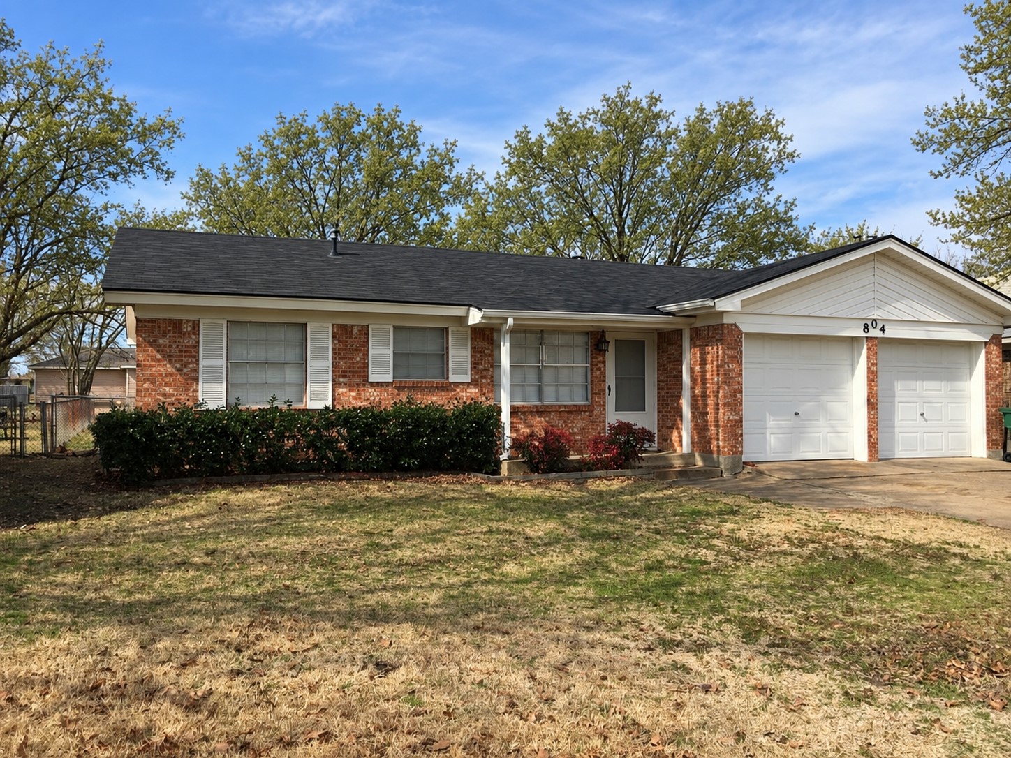 Single-story brick house with white siding, a two-car garage, and shrubs along the front.