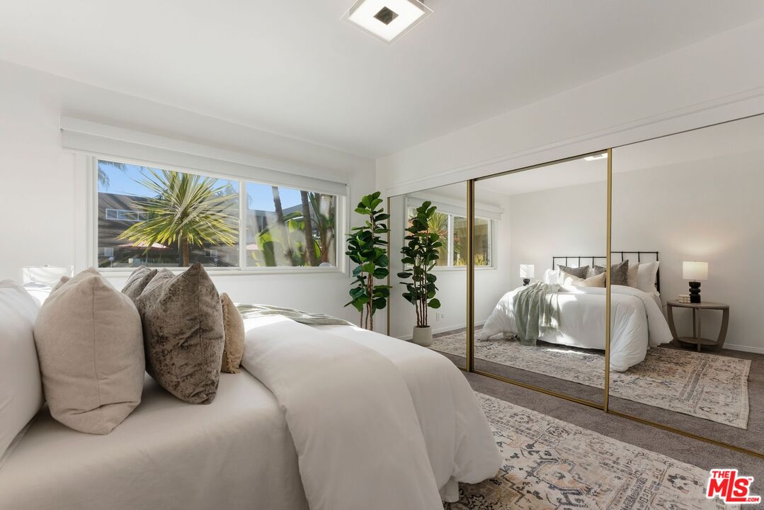 Staged bedroom with a white bed, mirrored closet doors, and a large window overlooking palm trees.