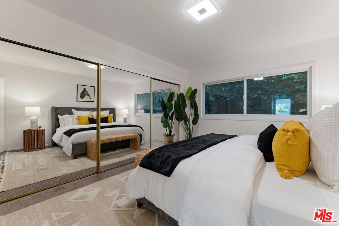 Staged bedroom with mirrored closet doors, mustard-yellow pillows, and large windows facing greenery.