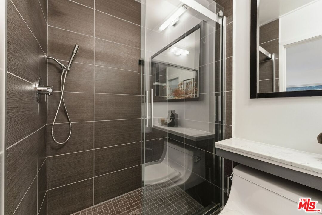 Bathroom with a glass-enclosed shower, dark tile, handheld showerhead, and white marble vanity.