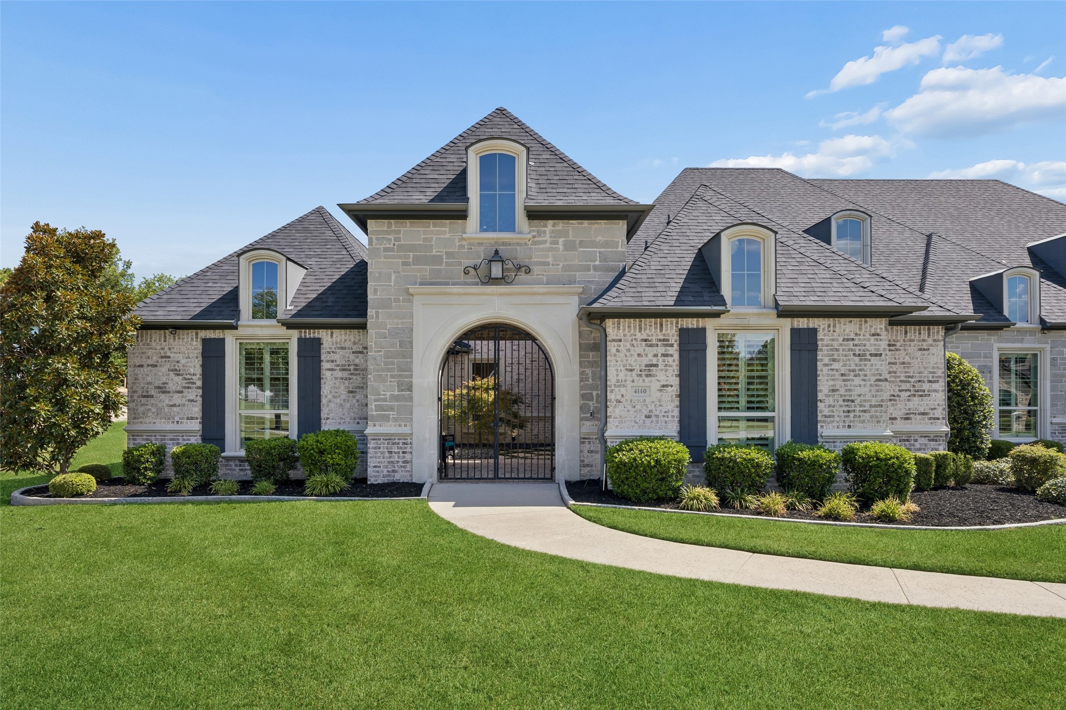 Gray stone-and-brick home with a central arched gated entrance and manicured lawn.