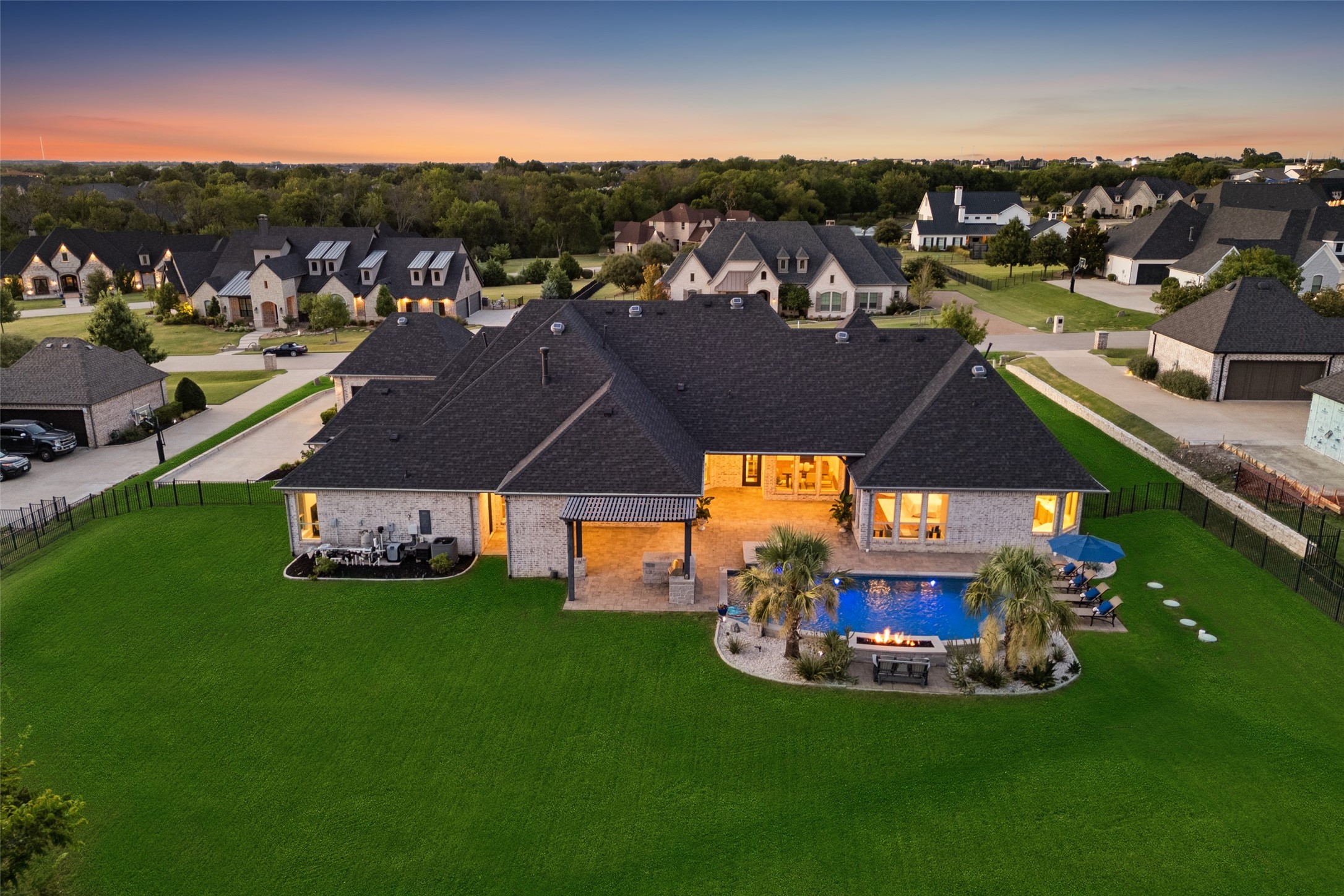 Aerial view of a brick home with a large lawn, covered patio, and illuminated pool at dusk.